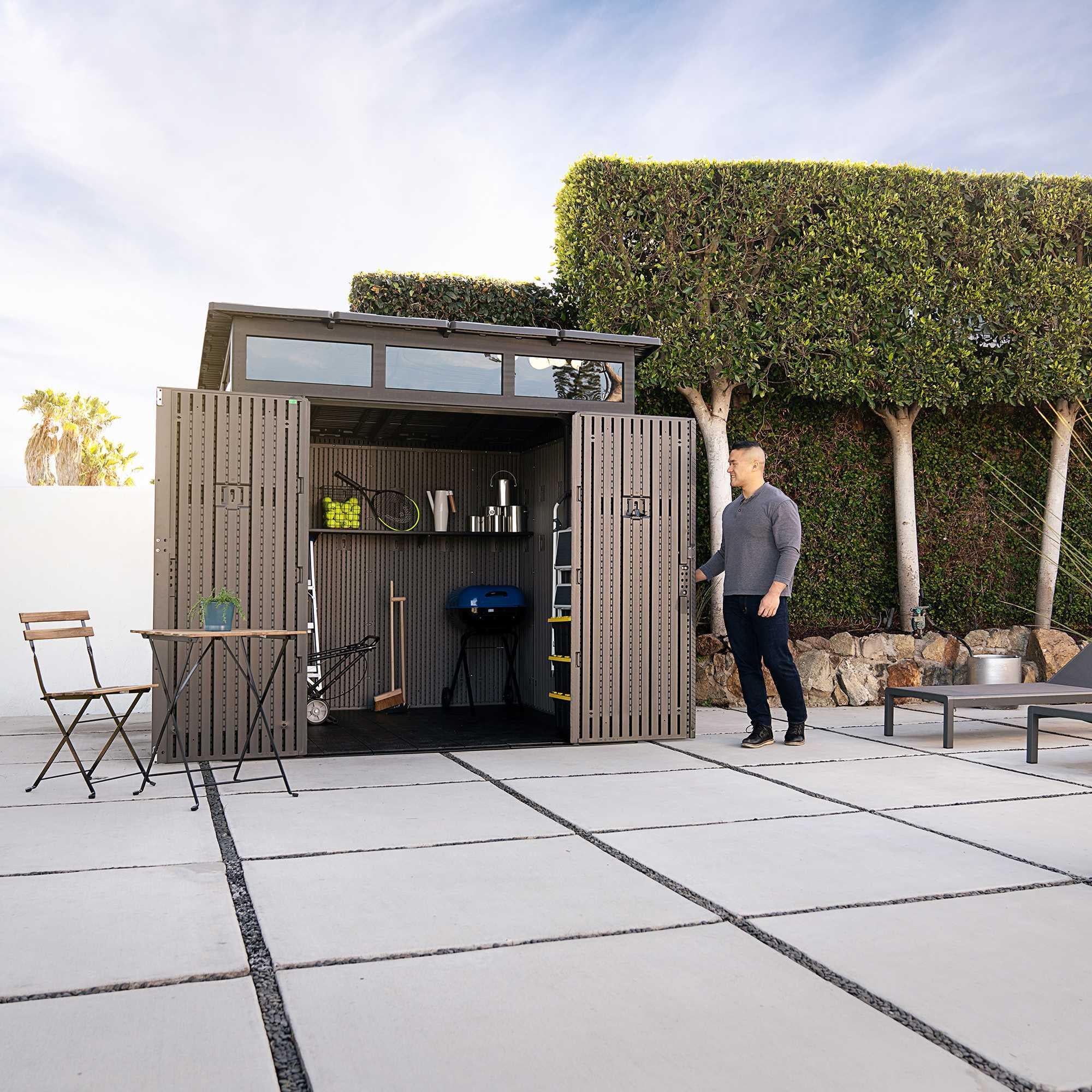 Man opening the double doors of a Lifetime Farmhouse Shed 60542 on a modern concrete patio, interior stocked with sports gear, grill, ladder, and shelved items